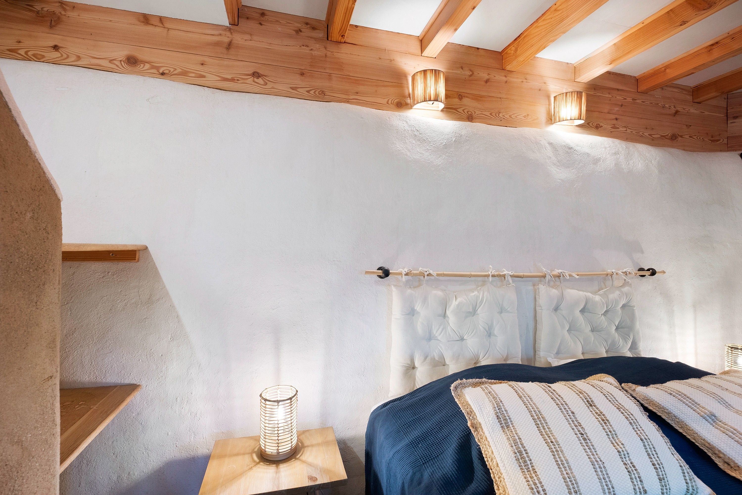 Ein Schlafzimmer mit geneigter Decke, Holzbalken, einem Bett mit Kissenmuster, einem Nachttisch mit Lampe und einer Gardinenstange mit Vorhang.