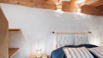 Ein Schlafzimmer mit geneigter Decke, Holzbalken, einem Bett mit Kissenmuster, einem Nachttisch mit Lampe und einer Gardinenstange mit Vorhang.