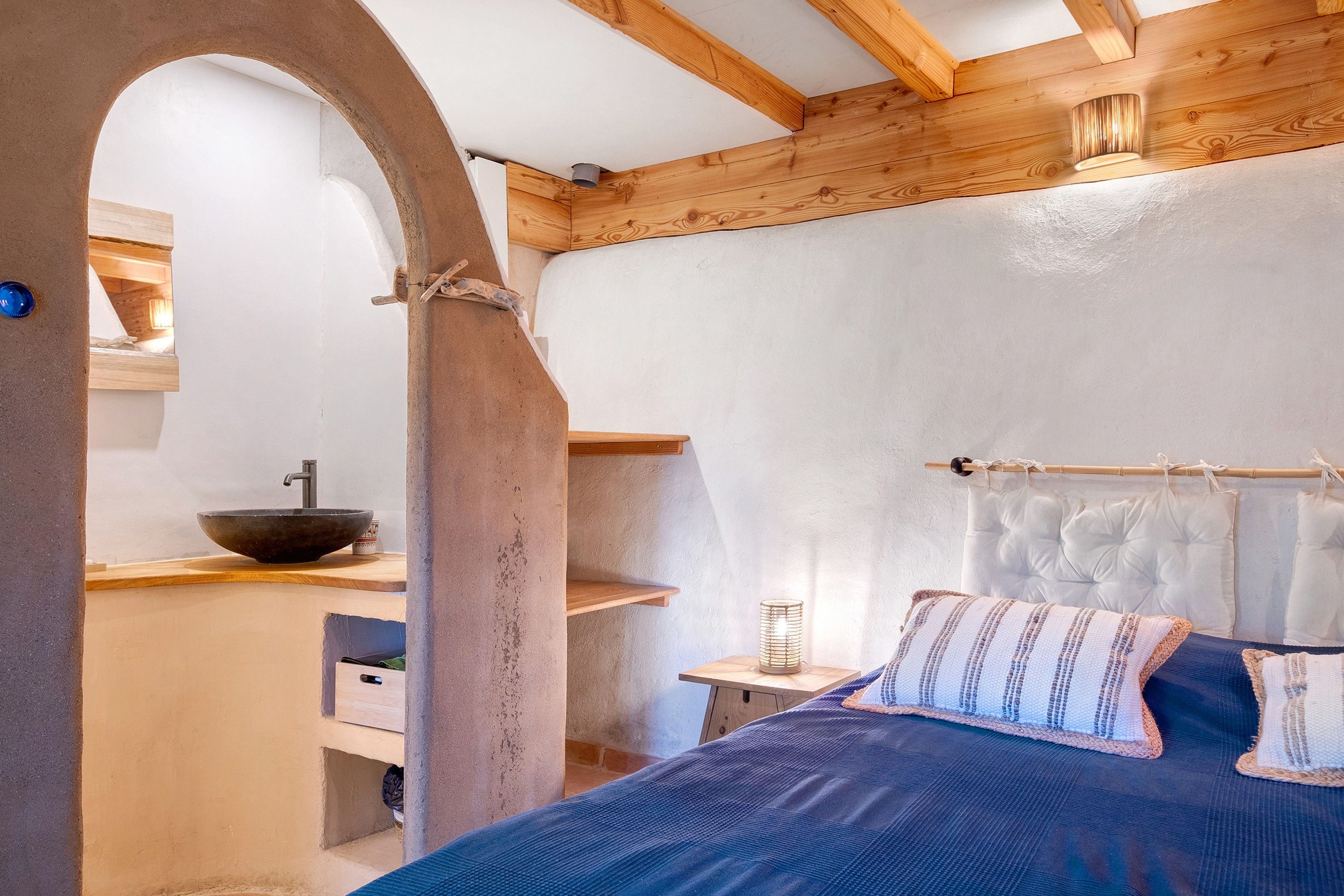 A bedroom with a bed, a wooden headboard, a bedside table, a nightstand, a wall-mounted shelf, and a window with natural light.