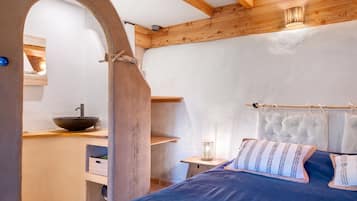 A bedroom with a bed, a wooden headboard, a bedside table, a nightstand, a wall-mounted shelf, and a window with natural light.