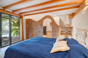 A bedroom with a large bed, wooden beams, a view of greenery, and a unique wall design.