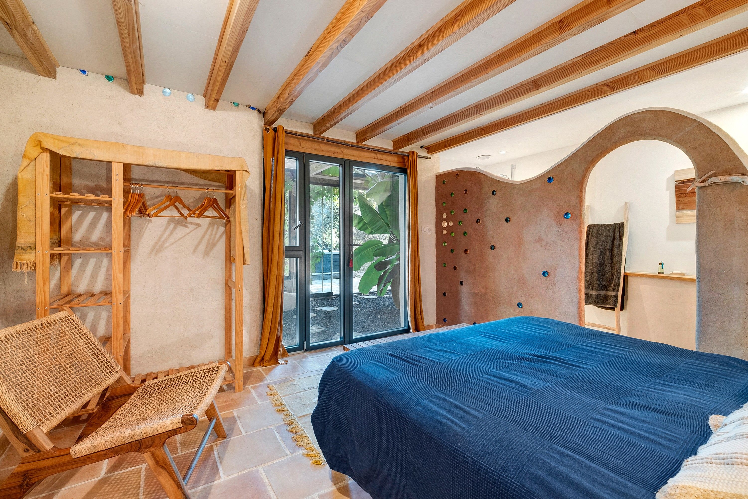 A bedroom with a bed, a wooden wardrobe, a wicker chair, and a sliding glass door leading to an outdoor area.