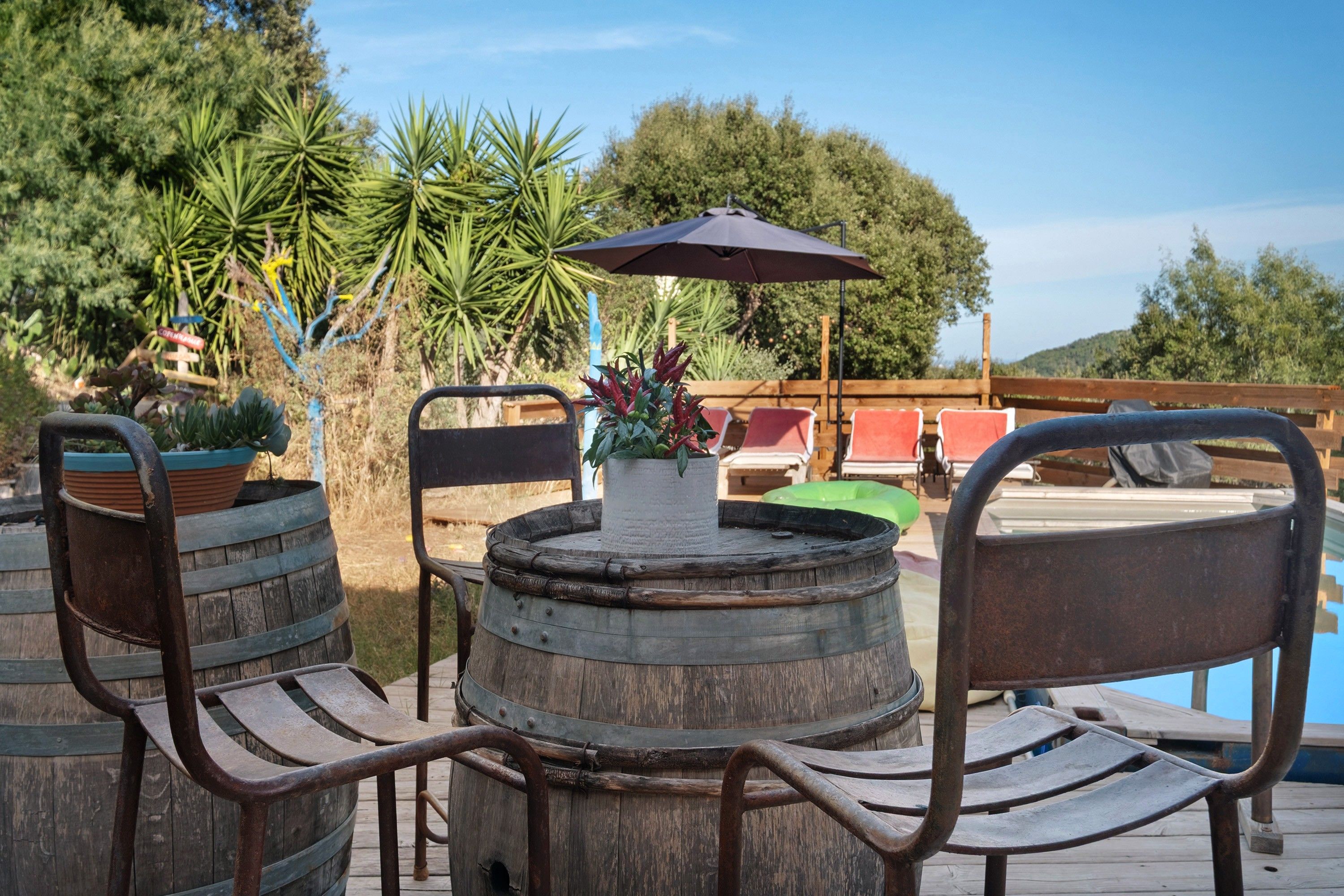 Une terrasse en bois avec une table ronde et des chaises, entourée de plantes en pot et d’un espace piscine.