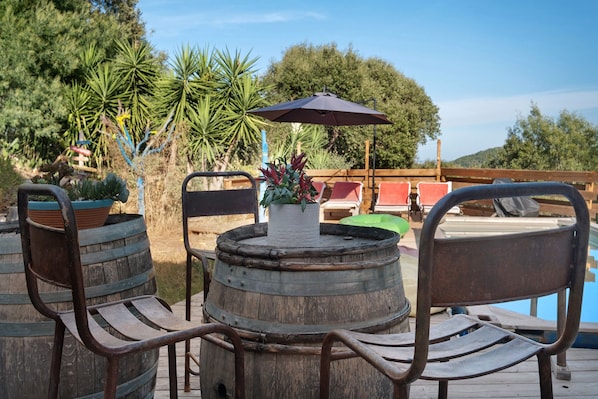 A wooden deck with a round table and chairs, surrounded by potted plants and a pool area.