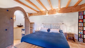 Une chambre avec un plafond en bois, un lit avec une literie bleue, une petite table et un mur orné d’une tapisserie colorée.