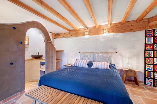 A bedroom with a wooden ceiling, a bed with blue bedding, a small table, and a wall with a colorful tapestry.
