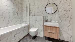 A bathroom with a marble wall, a bathtub, a toilet, a sink with a mirror, and a vanity cabinet. - Bank Polski City Center with Parking (Gdynia)