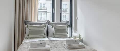 Una cama bien tendida con un edredón texturizado, dos almohadas con borlas y un juego de toallas dobladas sobre una mesita de noche.