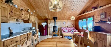 A wooden cabin interior with a central dining table, wooden cabinets, and a hanging light fixture.
