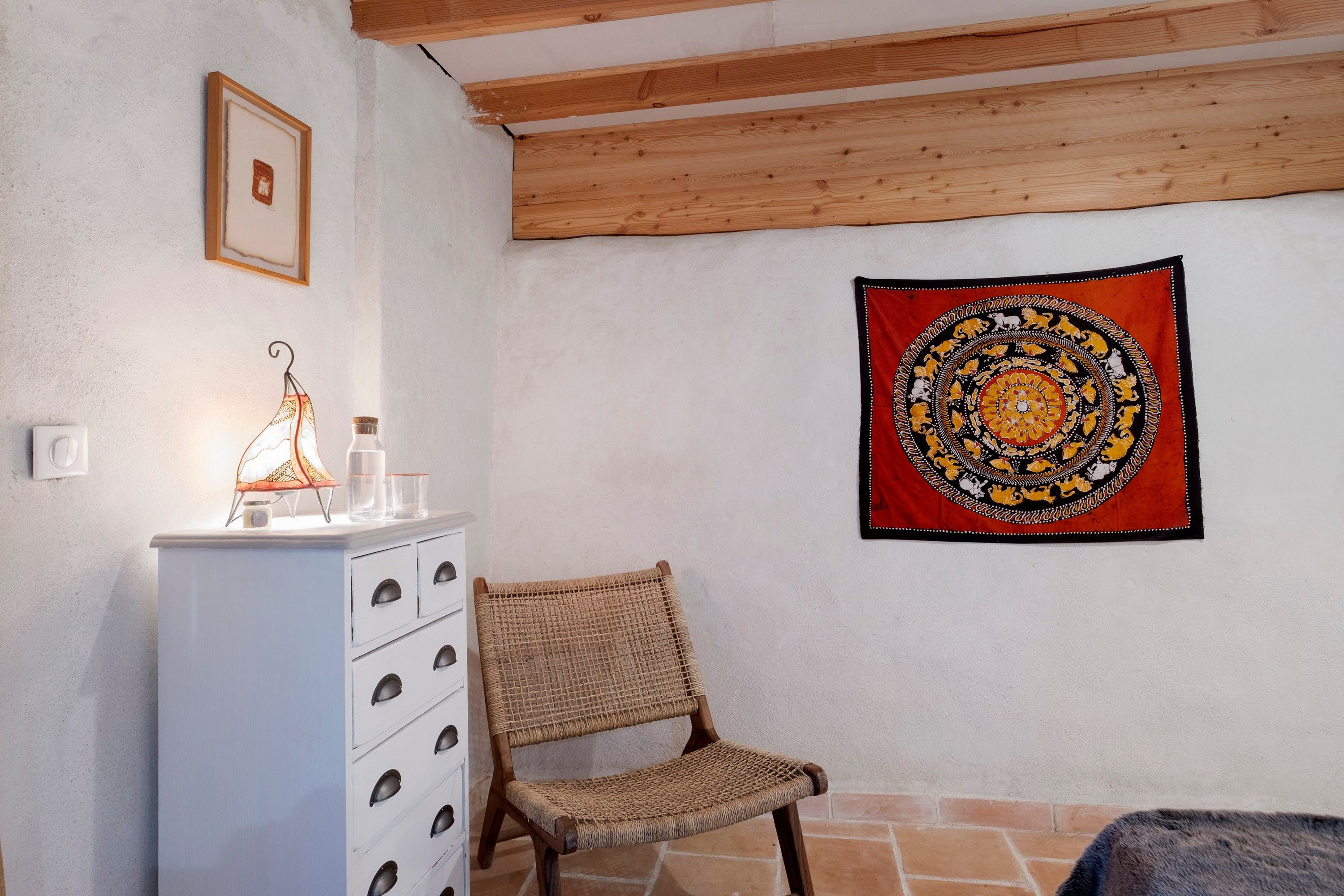 Une pièce avec une armoire blanche, une chaise en osier, une tapisserie et une poutre en bois au plafond.
