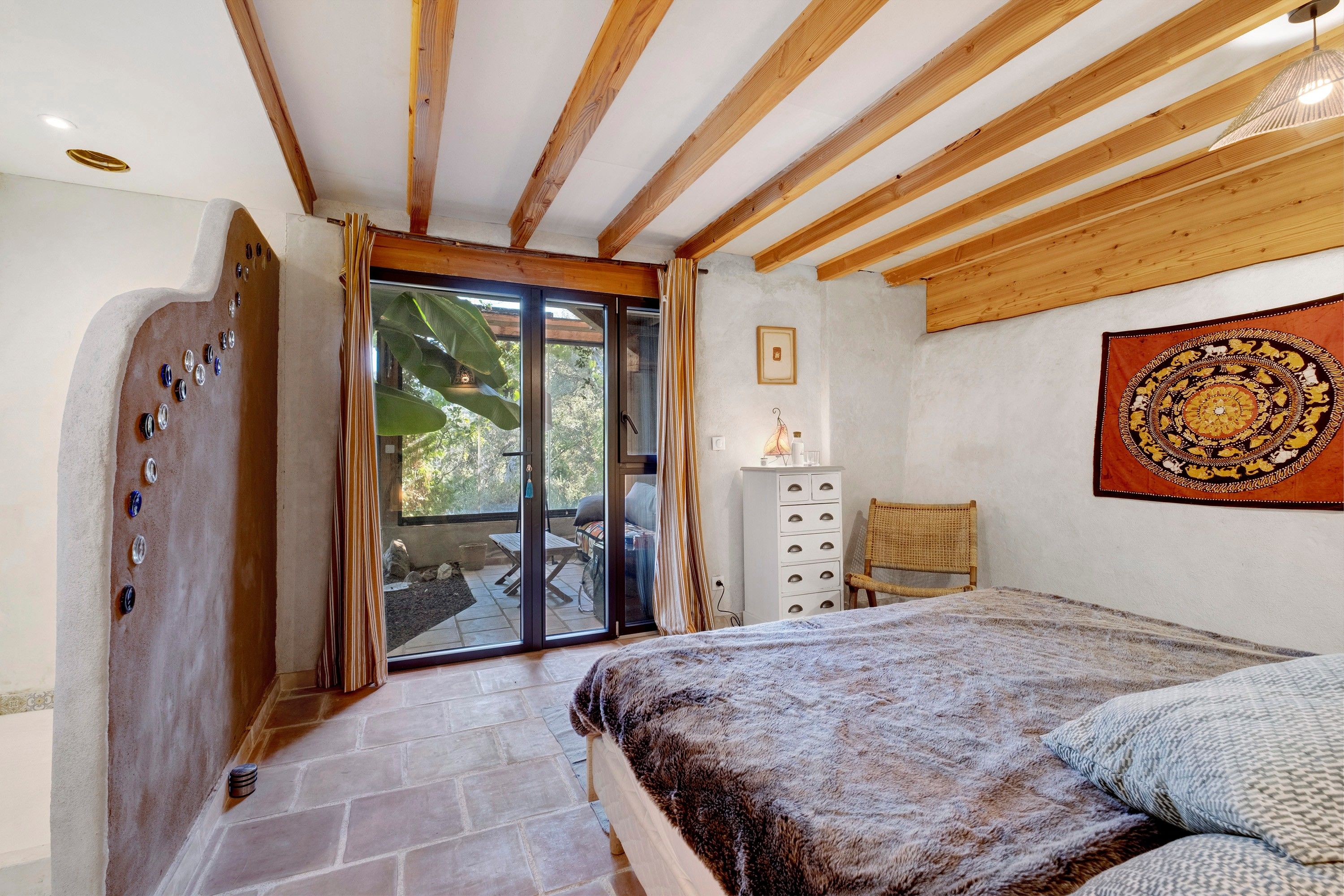 Een slaapkamer met een bed, een houten plafond, een schuifdeur met glas, een klein kastje en een wandtapijt.