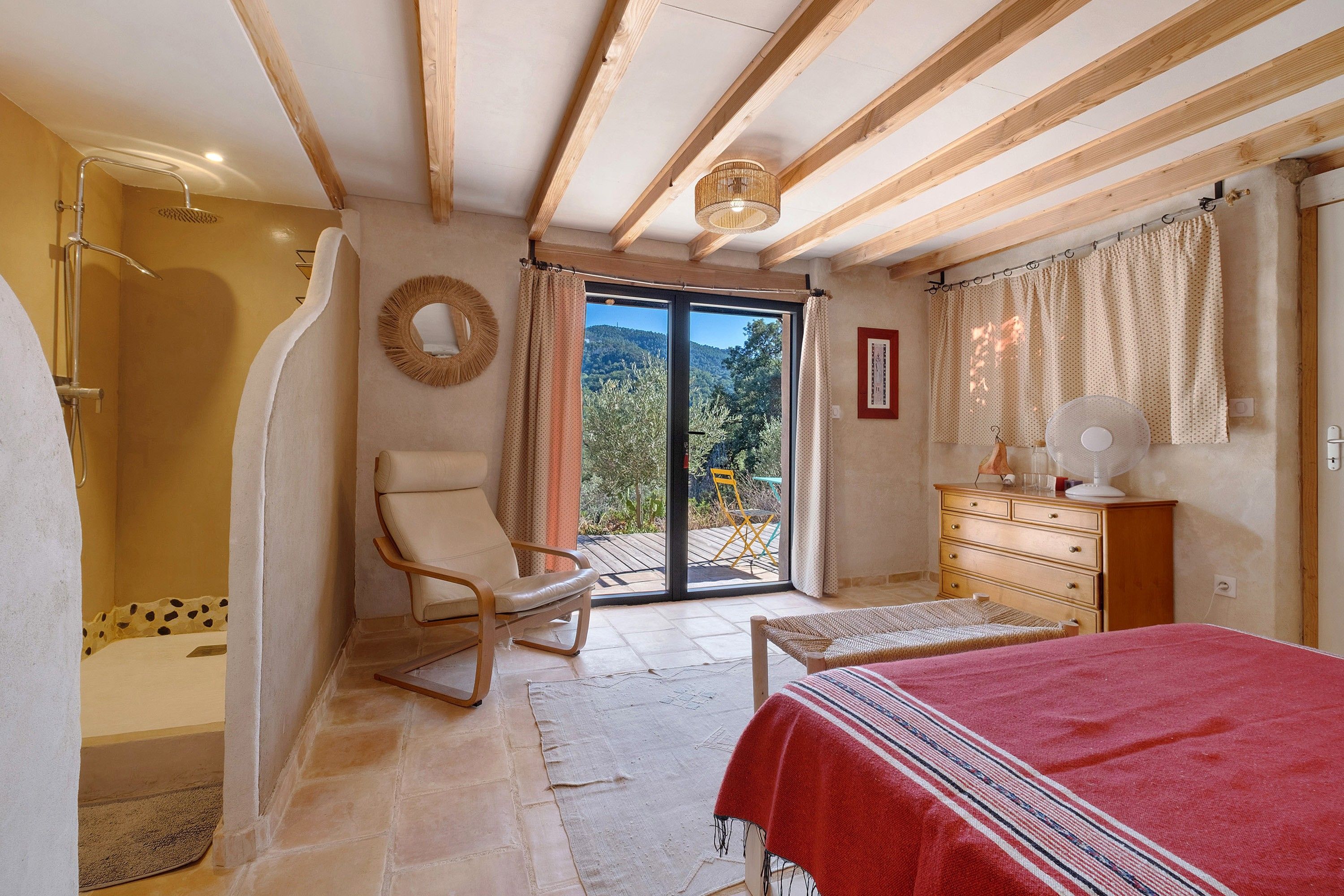Ein Schlafzimmer mit Bett, Sessel, Kommode, Ventilator und Blick ins Freie.