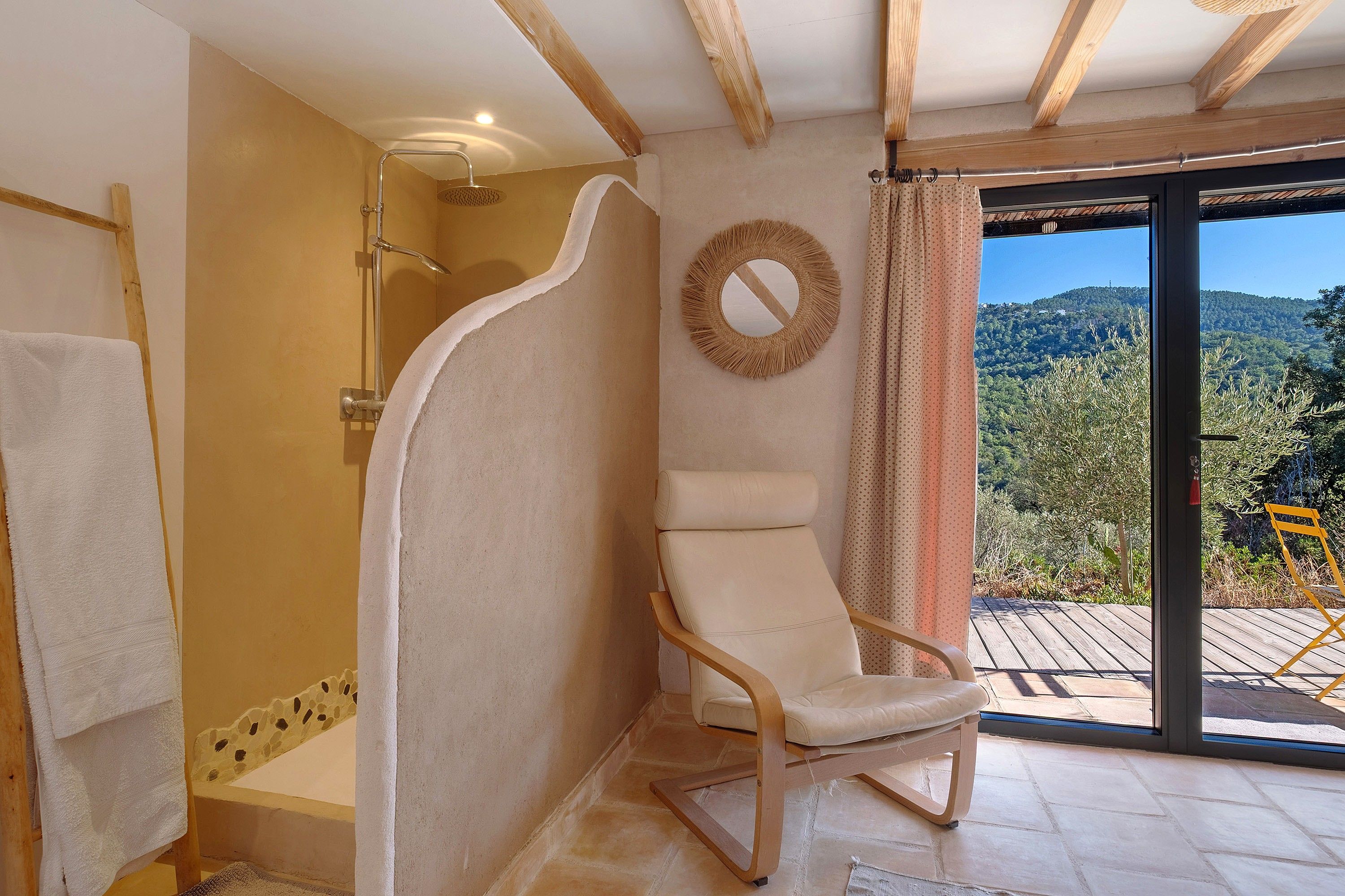 Une salle de bain avec une douche, une chaise, un miroir et une vue sur une forêt, visible à travers une porte vitrée.
