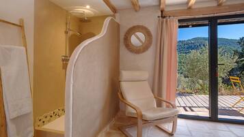 Une salle de bain avec une douche, une chaise, un miroir et une vue sur une forêt, visible à travers une porte vitrée.