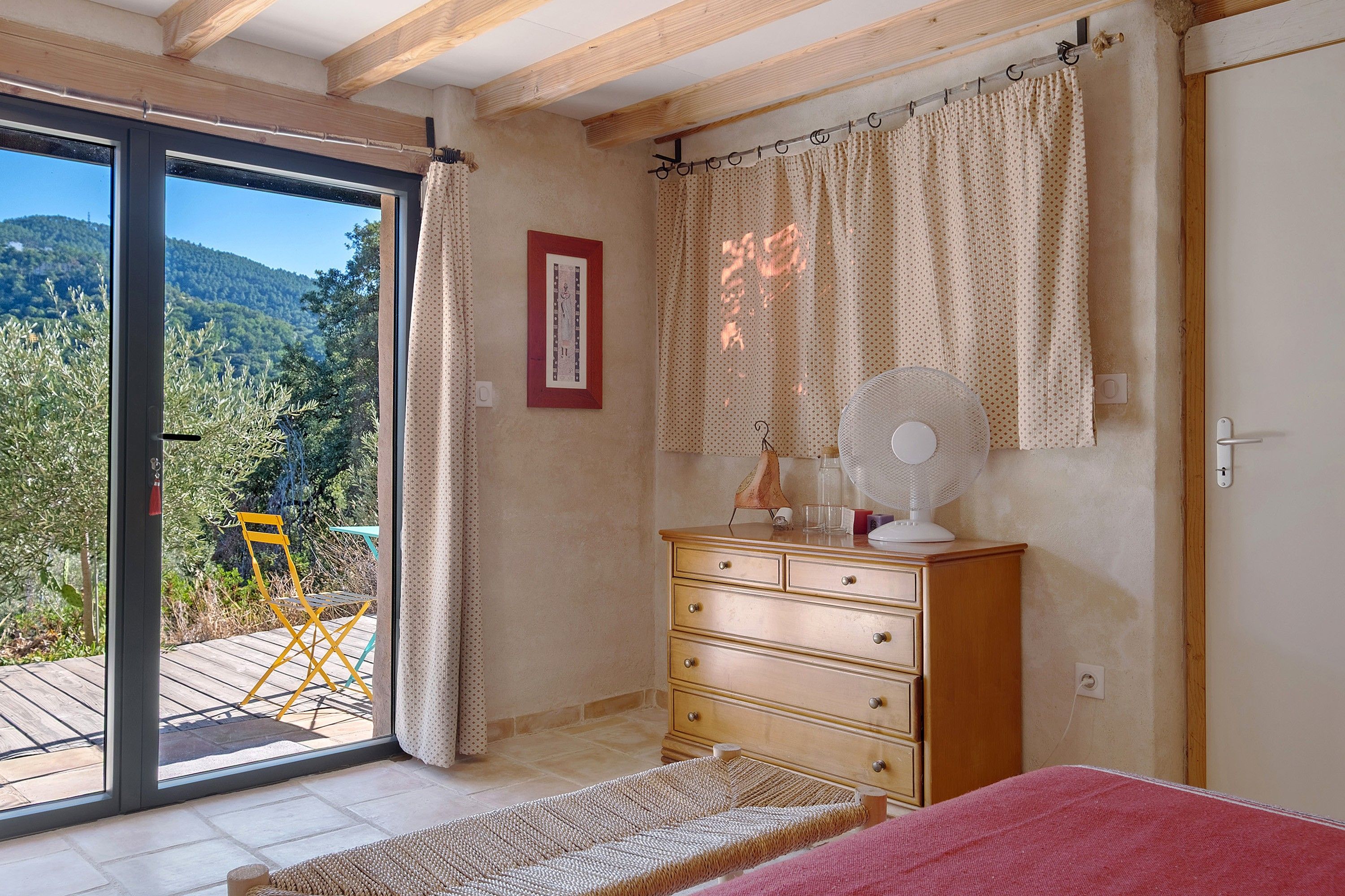 Een slaapkamer met een schuifglasdeur naar een buitenruimte, een houten dressoir, een bed en een ventilator bevestigd aan de muur.