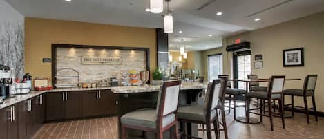 A hotel kitchen with a 'FREE DAILY BREAKFAST' sign, a buffet counter, and a dining area with chairs and tables.