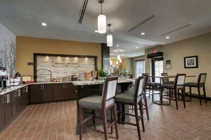 A hotel kitchen with a 'FREE DAILY BREAKFAST' sign, a buffet counter, and a dining area with chairs and tables.