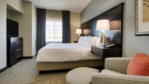 A hotel room with a large bed, bedside lamps, a desk with a telephone, a television, and a framed picture on the wall. - Hearing Accessible King Suite | Free Breakfast! (The Colony)