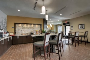 Una cocina de hotel con un letrero que dice 'DESAYUNO DIARIO GRATUITO', un mostrador de buffet y un comedor con sillas y mesas.
