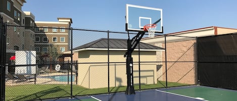 An outdoor basketball court with a chain-link fence, a basketball hoop, and a pool in the background.