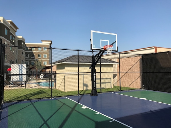 An outdoor basketball court with a chain-link fence, a basketball hoop, and a pool in the background. - ADA King Studio w/ Roll-In Shower | Free Breakfast (The Colony)
