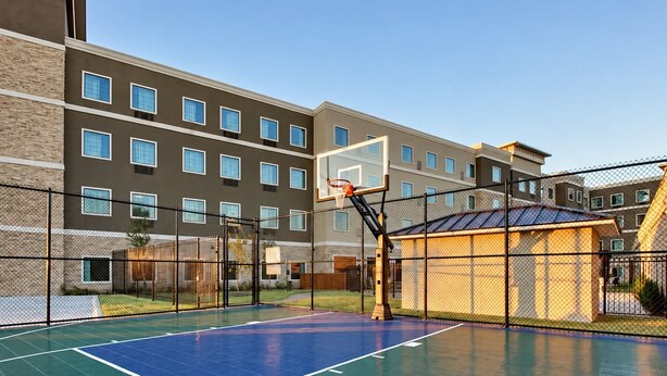 An outdoor basketball court with a basketball hoop, surrounded by a fence and a building with multiple windows. - Accessible 1-Bdrm Suite w/ 2 Queens & Pool (The Colony)