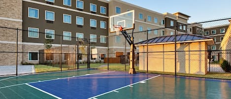 An outdoor basketball court with a basketball hoop, surrounded by a fence and a building with multiple windows.