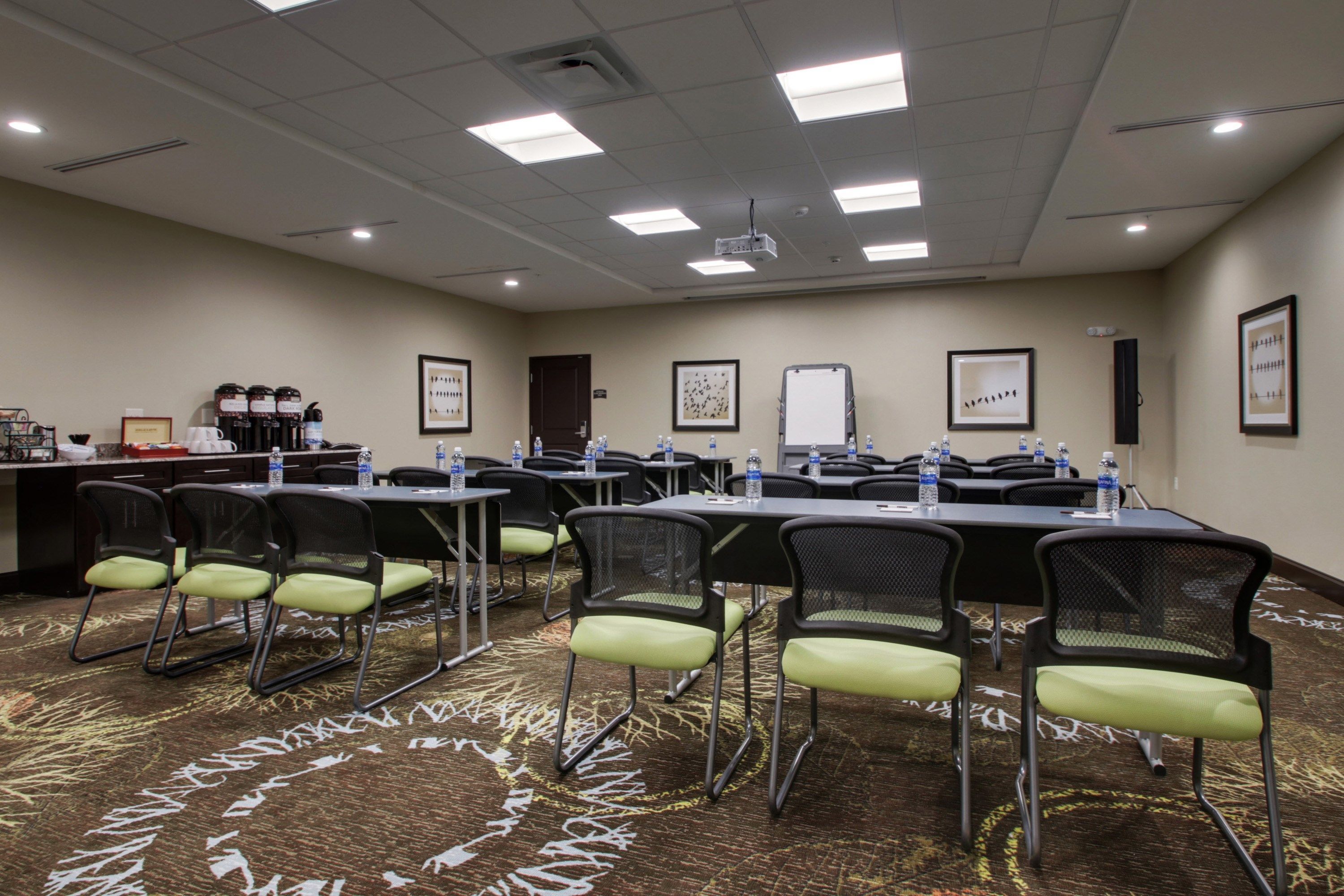 A conference room with tables and chairs, water bottles, framed pictures, and a projector on the ceiling.
