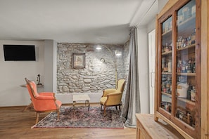 A living room with a stone wall, two chairs, a small table, a wooden cabinet with glass doors, and a television mounted on the wall.