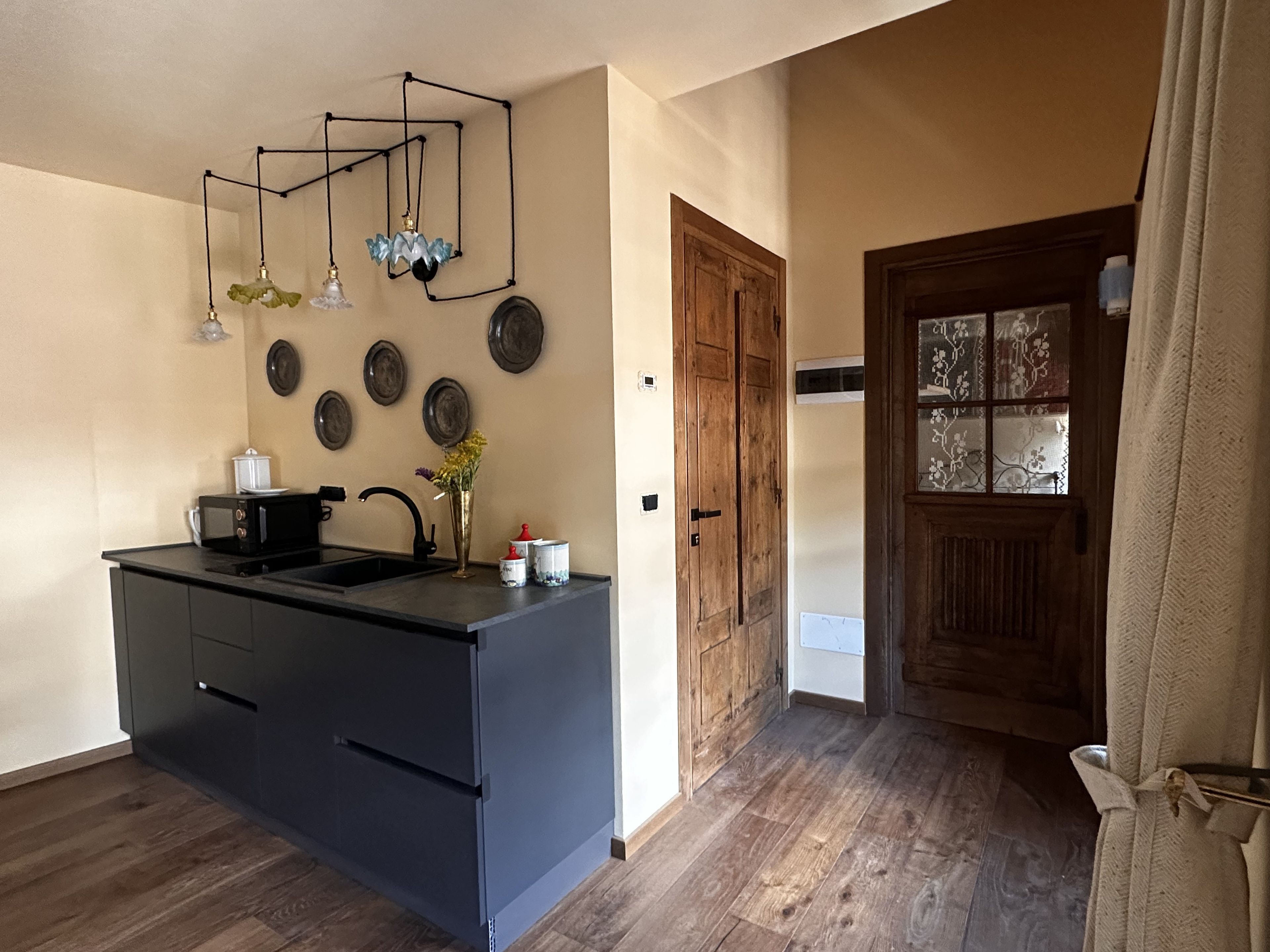 Eine Küche mit schwarzer Arbeitsplatte, einer Spüle, einer Mikrowelle und einer Vase mit Blumen. An der Wand hängen Teller und im Hintergrund ist eine Holztür zu sehen.
