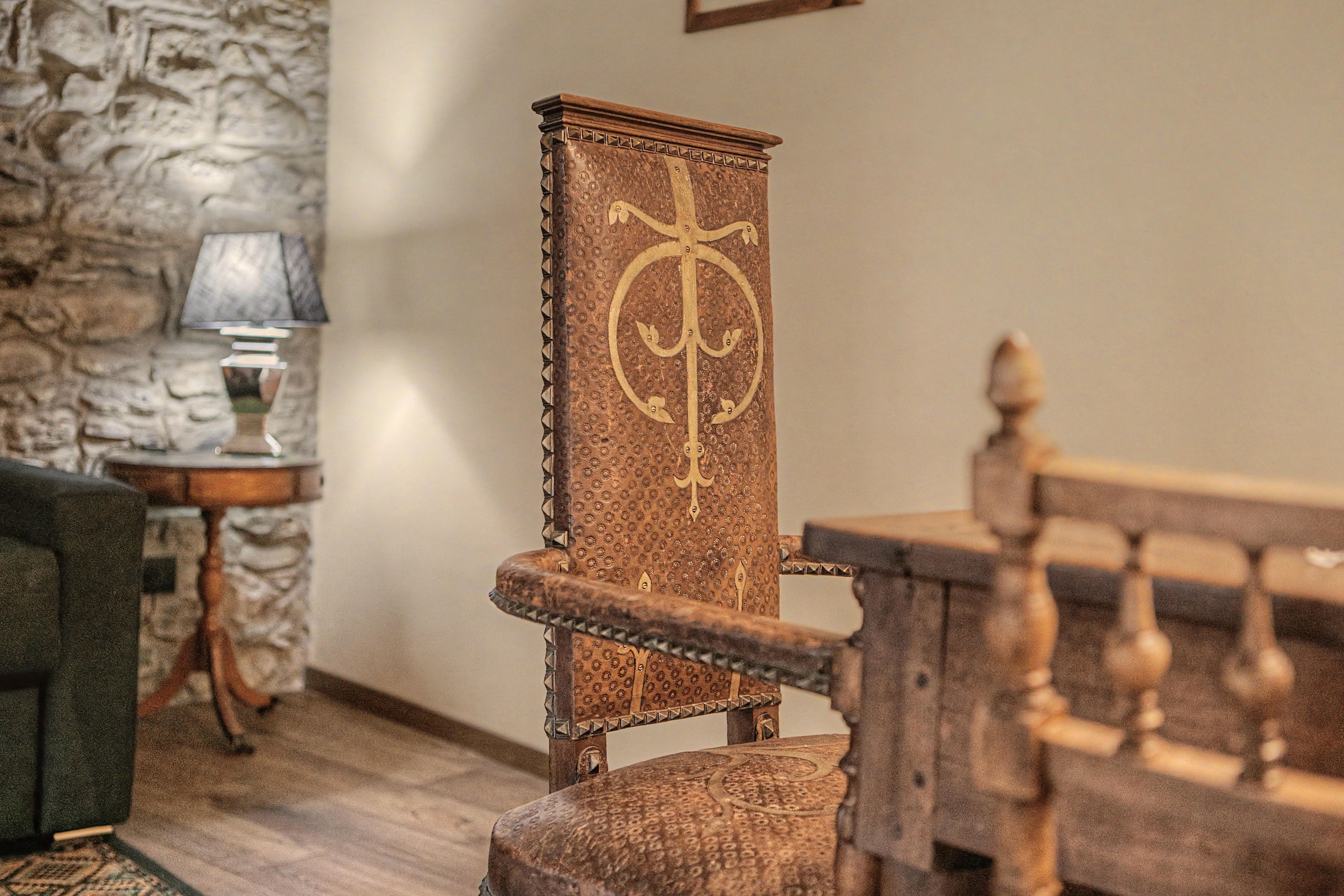 Une chaise en bois ornée d’un emblème en forme de croix, un mur de pierre et une lampe posée sur une table d’appoint.