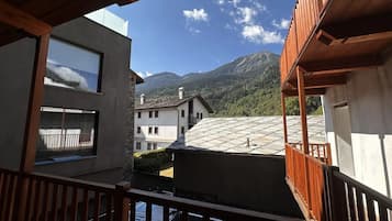 A view from a balcony with a mountain backdrop, featuring a building with a sloped roof and a wooden railing.