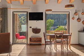 A cozy living room with a wooden ceiling, a TV mounted on the wall, and a balcony with a view of trees and buildings.