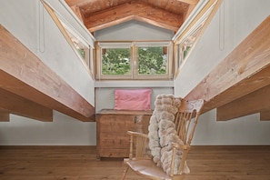 Una habitación con techo inclinado, vigas de madera, una ventana con vistas a la vegetación, un cofre de madera y un sillón con un plaid de piel.