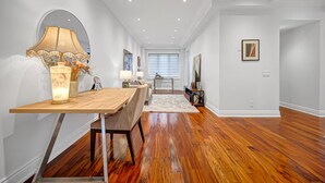 Interior moderno com mesa de jantar de madeira, luminária e vista para uma sala de estar com sofá e televisão.