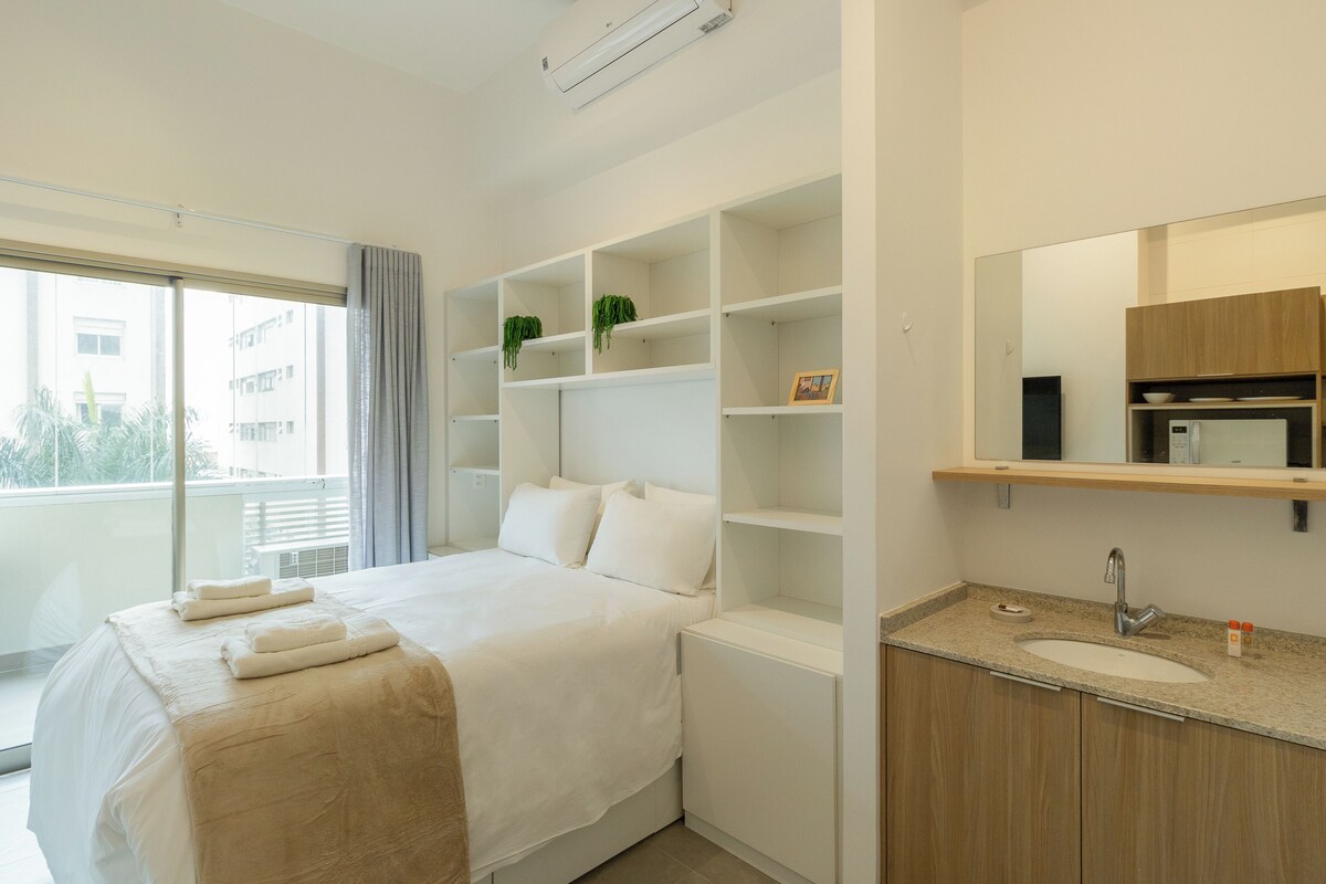 A hotel room with a bed, a built-in shelf with plants, a bathroom with a sink and mirror, and a window with a view of buildings.