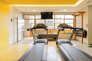 A gym with two treadmills, a television above them, and a view of buildings through large windows. - E-side By Bhomy (São Paulo)