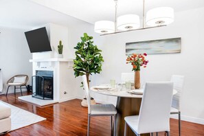 Un espace repas moderne avec une table ronde, des chaises blanches, une plante en pot et un téléviseur à écran plat fixé au-dessus de la cheminée.
