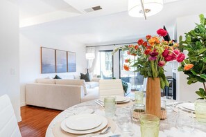 A modern living room with a dining area, featuring a white sofa, a dining table set with plates and glasses, and a vase with flowers.