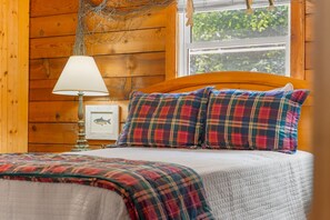 A wooden cabin interior with a bed, plaid bedding, a lamp, and a framed fish picture. - Gee’s Getaway is a cozy mountain retreat nestled in a peaceful setting just minutes from Boone. Relax by the fire, unwind in nature, and enjoy easy access to hiking, dining, shopping, and all the adventures the High Country has to offer. (Boone)