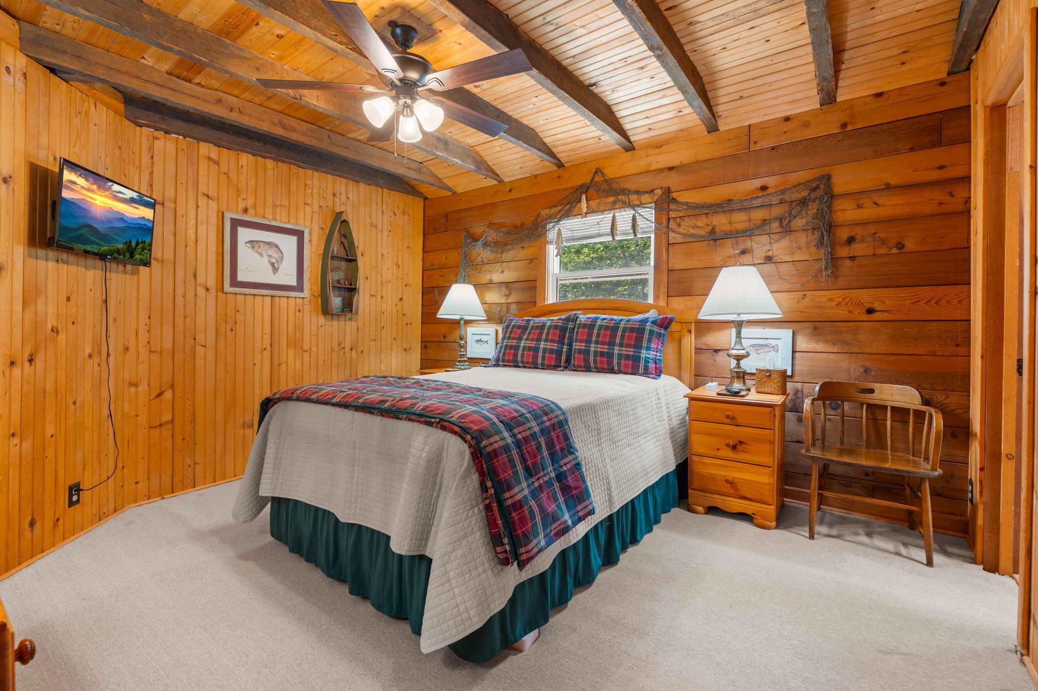 A wooden cabin bedroom with a bed, two bedside tables, a chair, a ceiling fan, and a window.