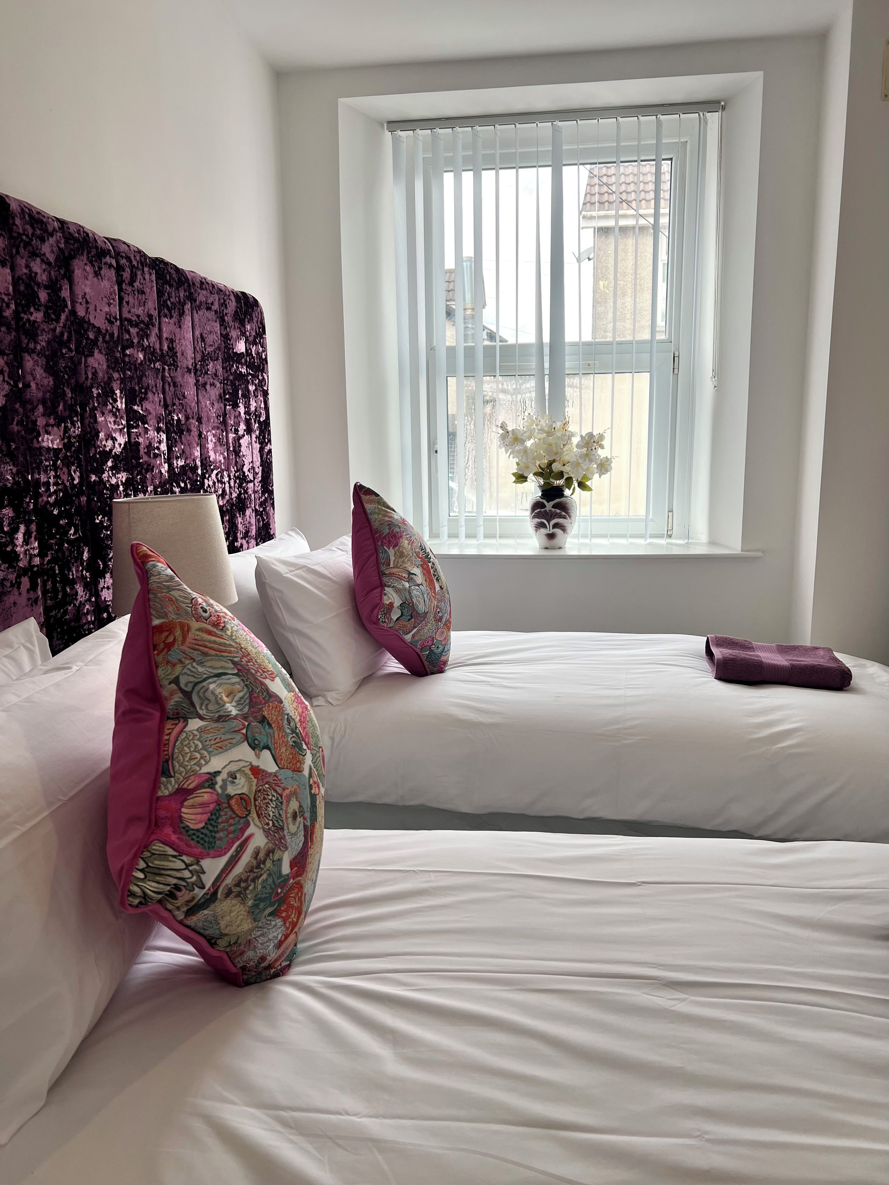 A bedroom with two beds, a window with blinds, a vase with flowers, and a headboard with a textured fabric.