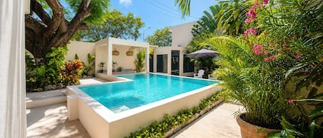 A rectangular swimming pool with a white border, surrounded by a variety of plants and trees, and a clear blue sky above.