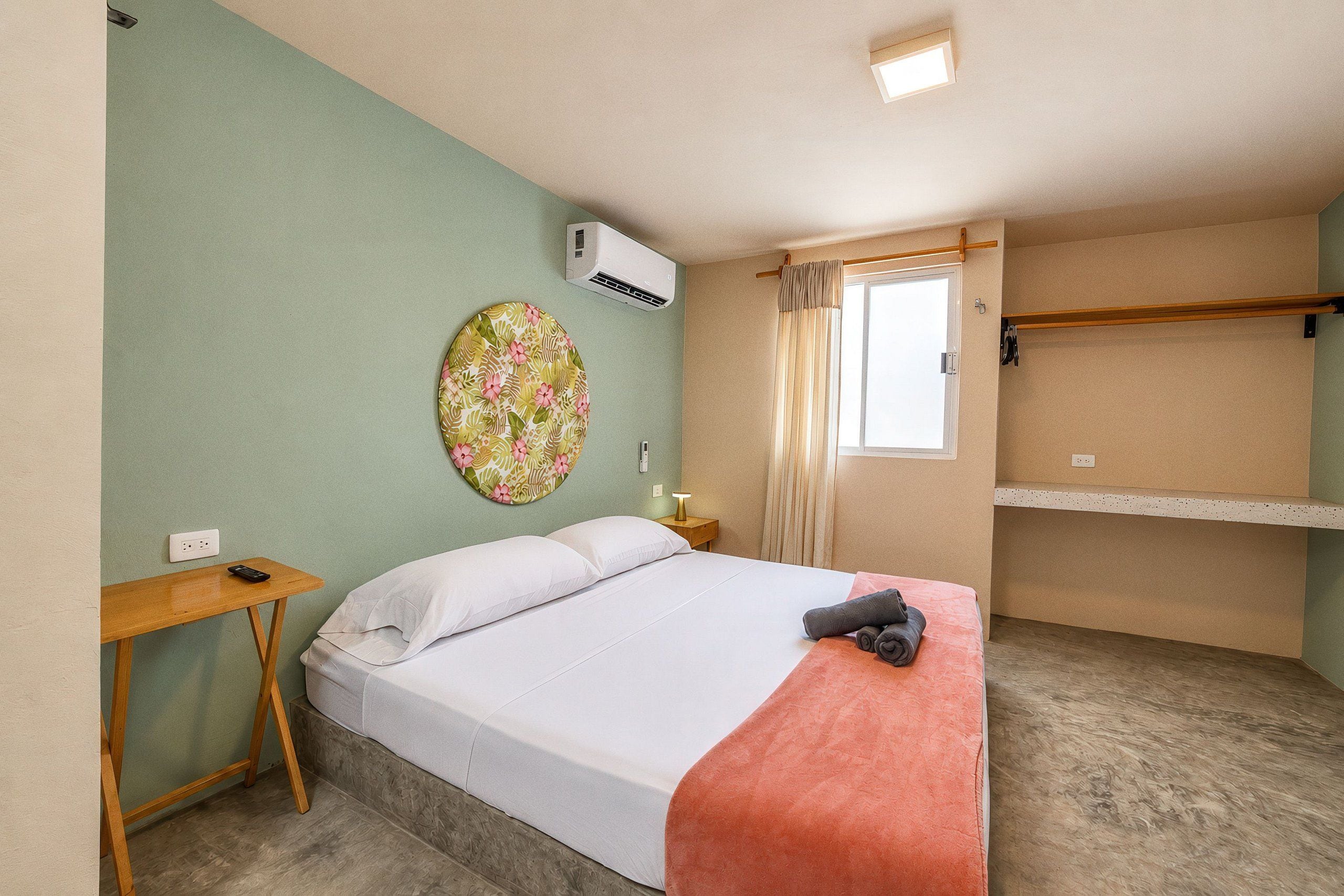 Una camera d'albergo con un letto, un comodino, un condizionatore d'aria a parete, una finestra con tende e un piccolo ripiano.