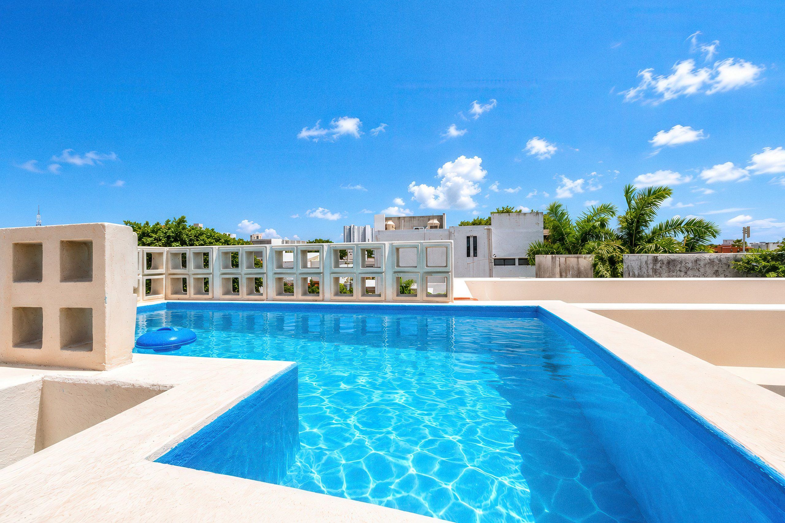 Piscina com água azul cristalina, rodeada por um deck de concreto e um edifício branco com várias janelas.