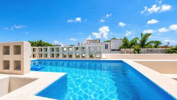 Piscina com água azul cristalina, rodeada por um deck de concreto e um edifício branco com várias janelas.