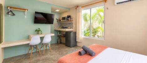 A compact hotel room with a bed, a desk with two chairs, a television, a small kitchenette, and a window with a view of greenery.