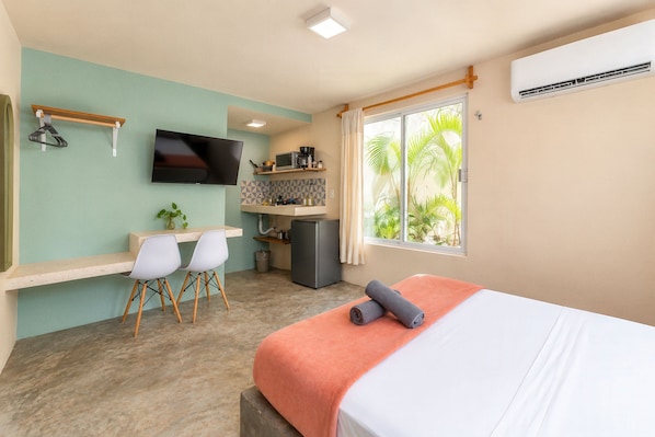 Habitación de hotel compacta con cama, escritorio con dos sillas, televisor, kitchenette pequeña y ventana con vista a vegetación.