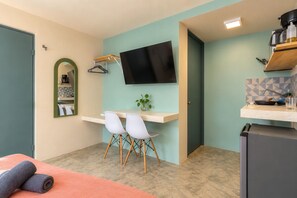 Una habitación compacta con kitchenette, cama con toallas enrolladas, escritorio con sillas, televisor de pantalla plana y espejo.