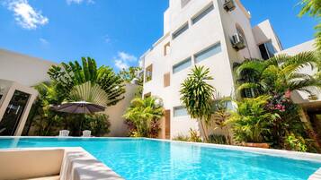 A swimming pool with a clear blue water surface, surrounded by lush tropical plants and a modern building with multiple windows.